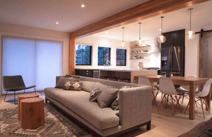 Modern open-concept living and kitchen area with a gray sofa, patterned rug, wood dining table, and pendant lights, featuring large windows and minimalist decor—perfect for relaxing after a day at Sasquatch Mountain Resort.
