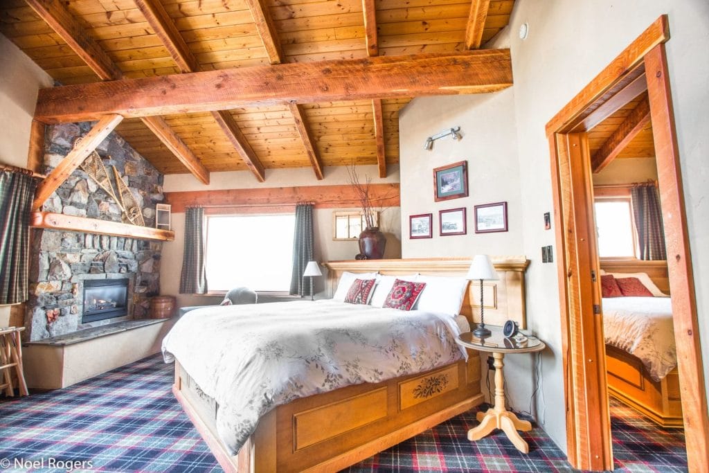 A cozy bedroom at Sasquatch Mountain Resort with a large bed, stone fireplace, wood-beam ceiling, plaid carpet, framed photos on the wall, and an open door leading to another room.