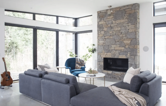 Modern living room with large windows, a stone fireplace, gray sectional sofa, blue armchair, white round tables, and a guitar in the corner—perfect for those seeking luxury Airbnb's in Whistler.