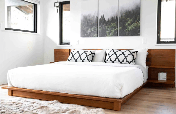 A modern bedroom with a low wooden bed frame, white bedding, patterned pillows, and forest-themed art above the bed—similar in style to luxury Airbnbs in Whistler. Natural light streams through two windows.
