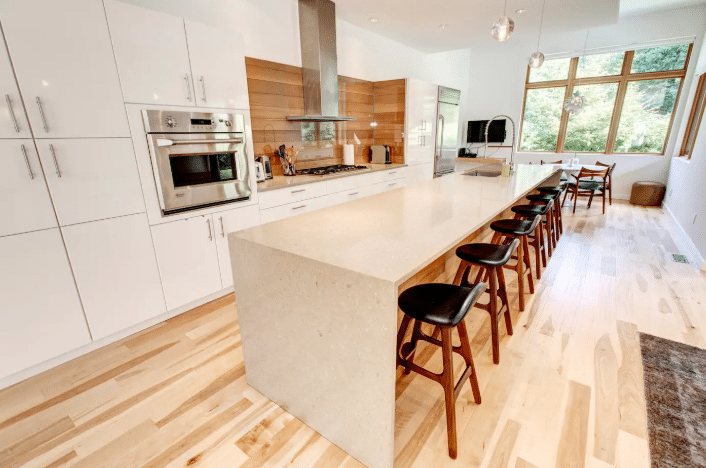 Modern kitchen with white cabinets, built-in stainless appliances, a long island with bar stools, wooden floor, and large windows letting in natural light—perfect for those seeking luxury Airbnb's in Whistler.