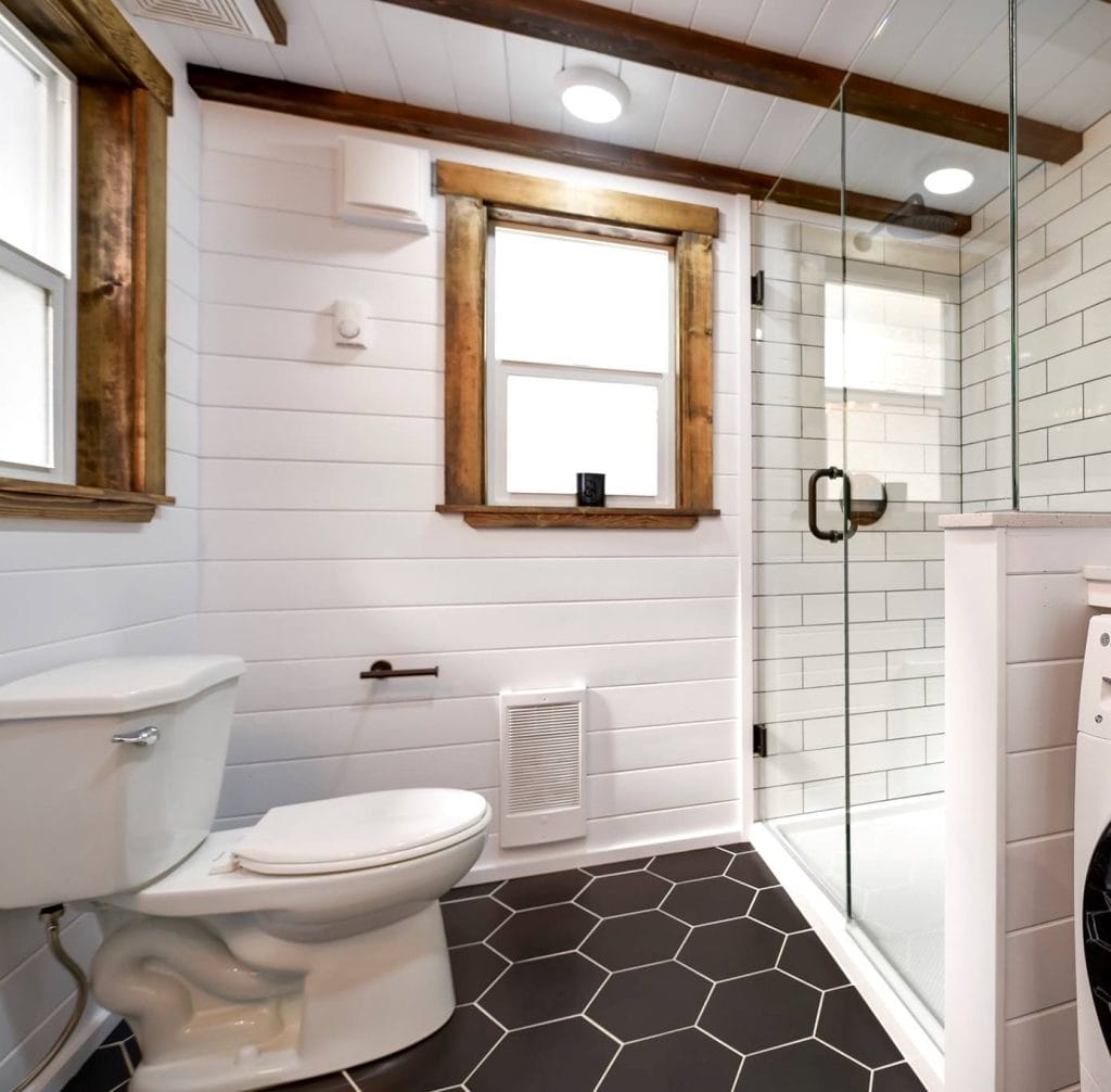 Modern bathroom in this BC Tiny House Airbnb features white shiplap walls, black hexagon floor tiles, a toilet, and a glass-enclosed subway-tiled shower. Two windows with wood trim fill the space with natural light.