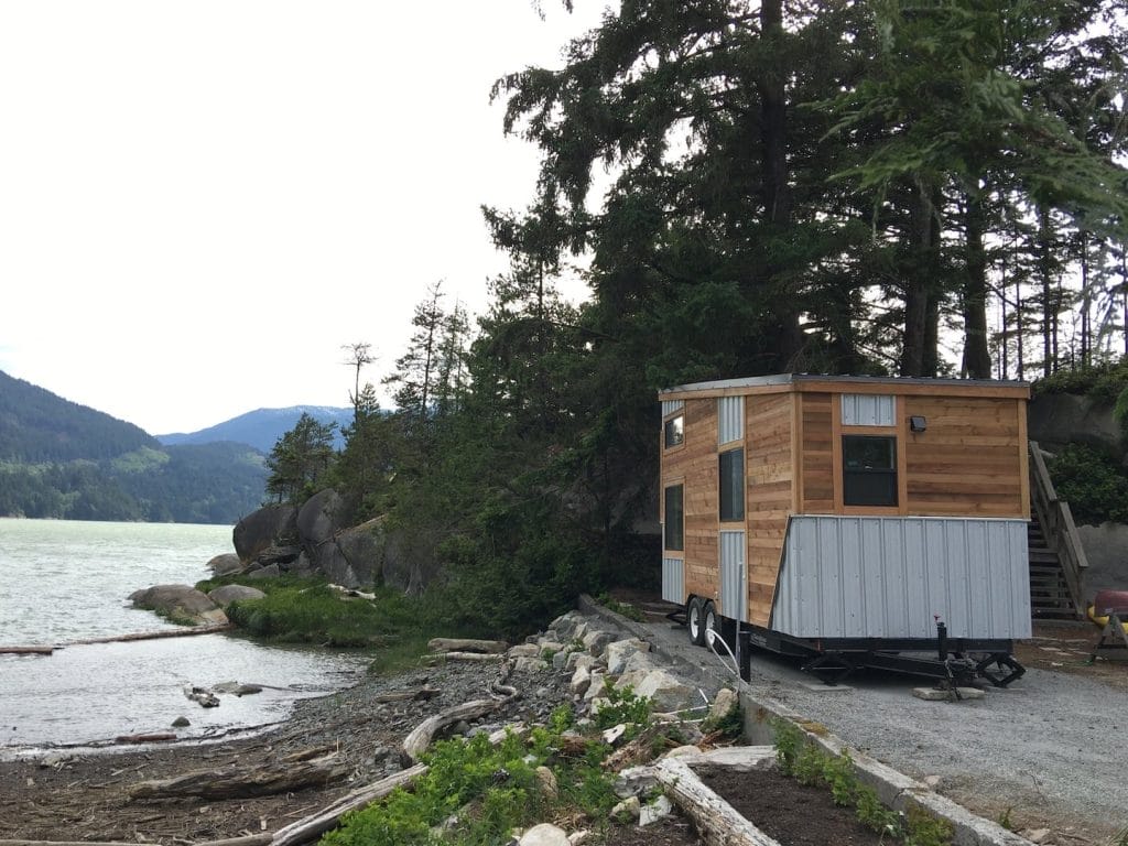 A small wooden BC Tiny House Airbnb on wheels is parked near a rocky shoreline, framed by trees, mountains, and sparkling water in the background.