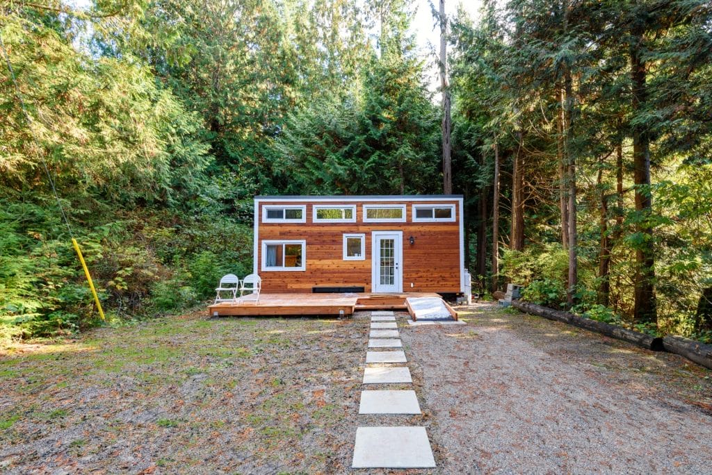 A small modern cabin with a wood exterior sits in a forest clearing, featuring a deck, two white chairs, and a stone path leading to the entrance—a cozy BC Tiny House Airbnb retreat.