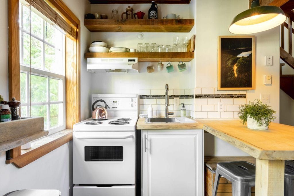 A small kitchen with a white stove, sink, open wooden shelves holding dishes, a window, potted plant on a wooden counter, and a metal stool—perfect for testing the best Airbnb cameras to capture cozy interiors.