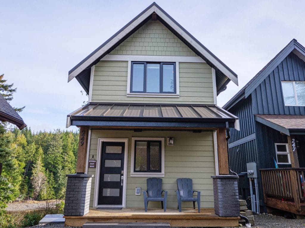 A two-story light green house with a covered front porch, two Adirondack chairs, modern black-trimmed windows and door—ideal for installing Best Airbnb Cameras for extra peace of mind.