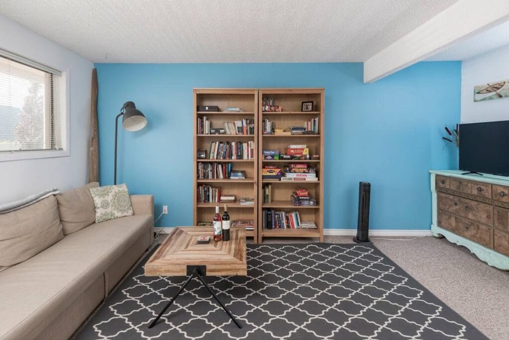 A living room with a beige sectional sofa, wooden coffee table, patterned rug, two bookshelves filled with books and games, and a TV on a teal dresser against a blue accent wall—perfect for relaxing after exploring the best Airbnb cameras.