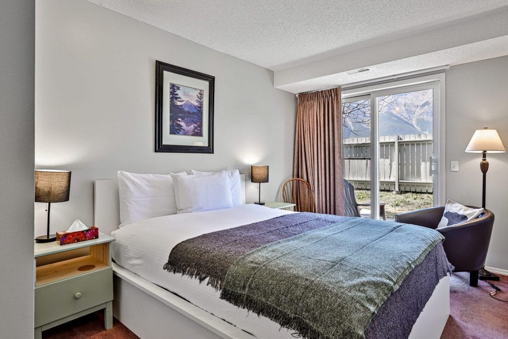 A bedroom with a neatly made bed, two bedside tables with lamps, an armchair by sliding glass doors, and a landscape painting on the wall—monitored discreetly for safety with one of the best Airbnb cameras.