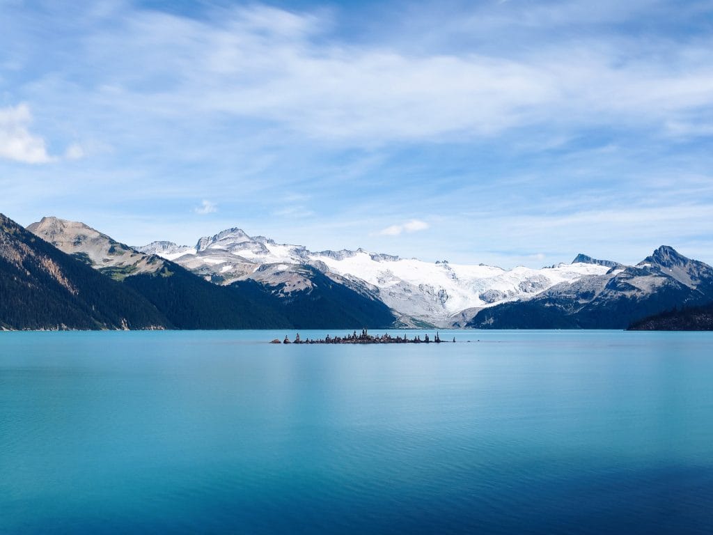 A calm turquoise lake with a small island in the center, surrounded by snow-capped mountains under a partly cloudy sky—a must-see among things to do in Whistler.