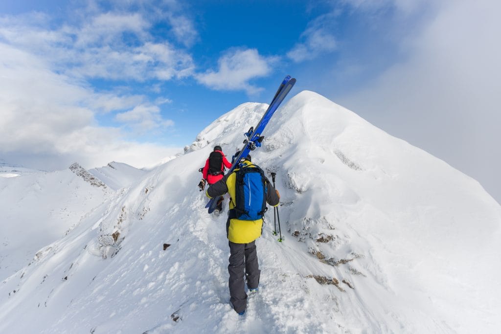 Two people in winter gear hike up a snow-covered mountain ridge near Banff ski resorts, carrying skis on their backpacks under a partly cloudy sky.