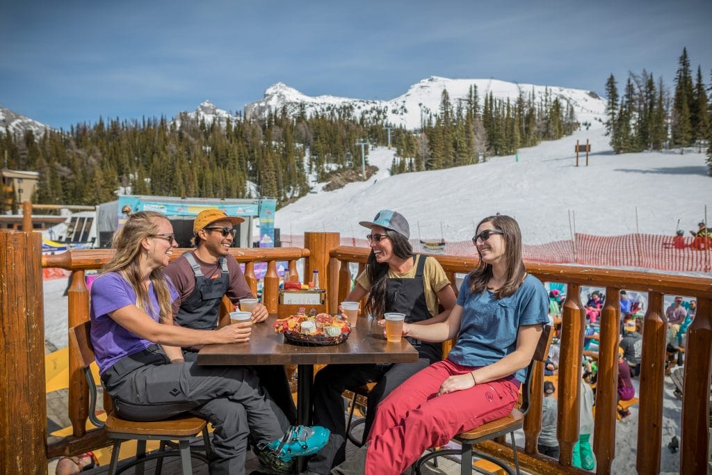 Four people sit at an outdoor table with drinks and snacks, smiling and talking, with the stunning snow-covered mountains and trees of Banff ski resorts in the background.
