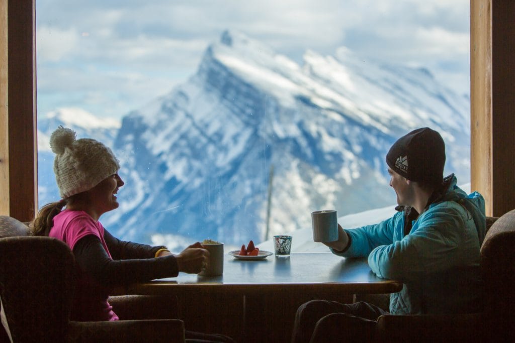 Two people sit at a table by a window with mugs, enjoying drinks and admiring the snowy mountain landscape of Banff ski resorts in the background.