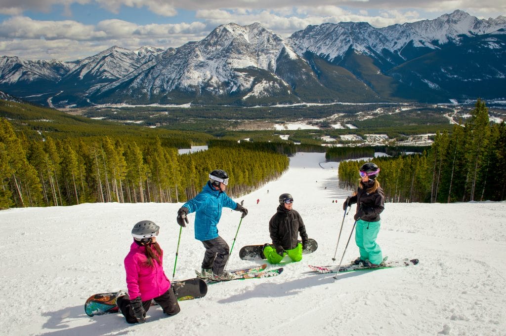 Four people in winter gear pause with skis and snowboards on a snowy mountain slope at one of the Banff ski resorts, surrounded by pine trees and snow-capped peaks.