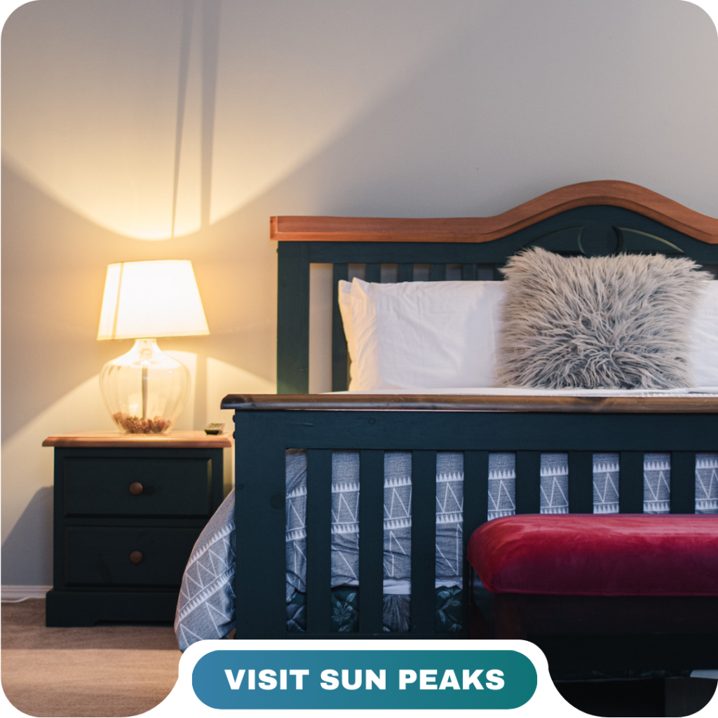 A neatly made bed with a gray fur pillow, wooden headboard, bedside table, and lamp. A red bench sits at the foot of the bed. Text reads "Visit Sun Peaks." Learn more about us and discover your perfect getaway.