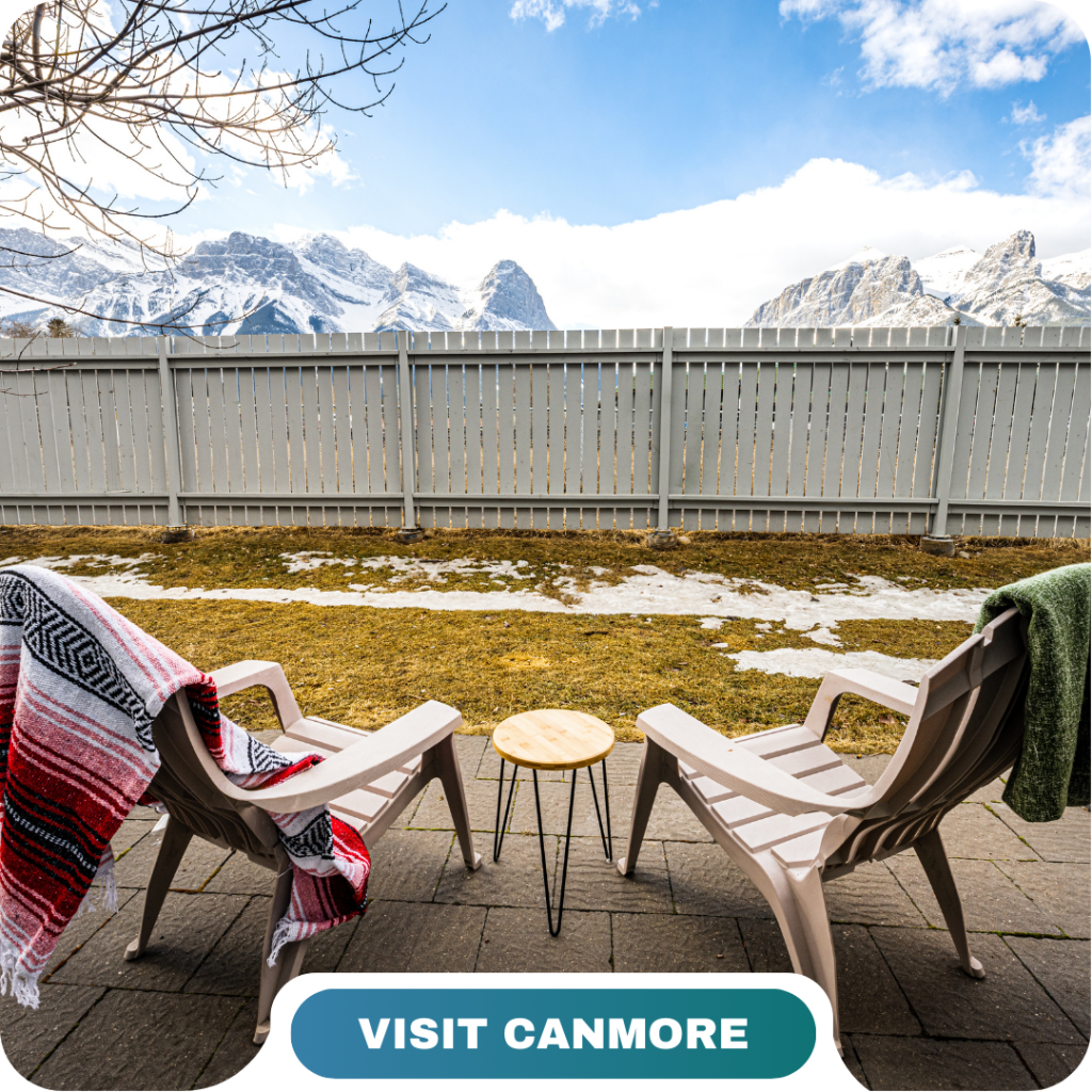 Two Adirondack chairs with blankets face a small table on a patio, overlooking a fenced yard with snow patches and snow-capped mountains in the background. Experience Canmore with Lifty Life.