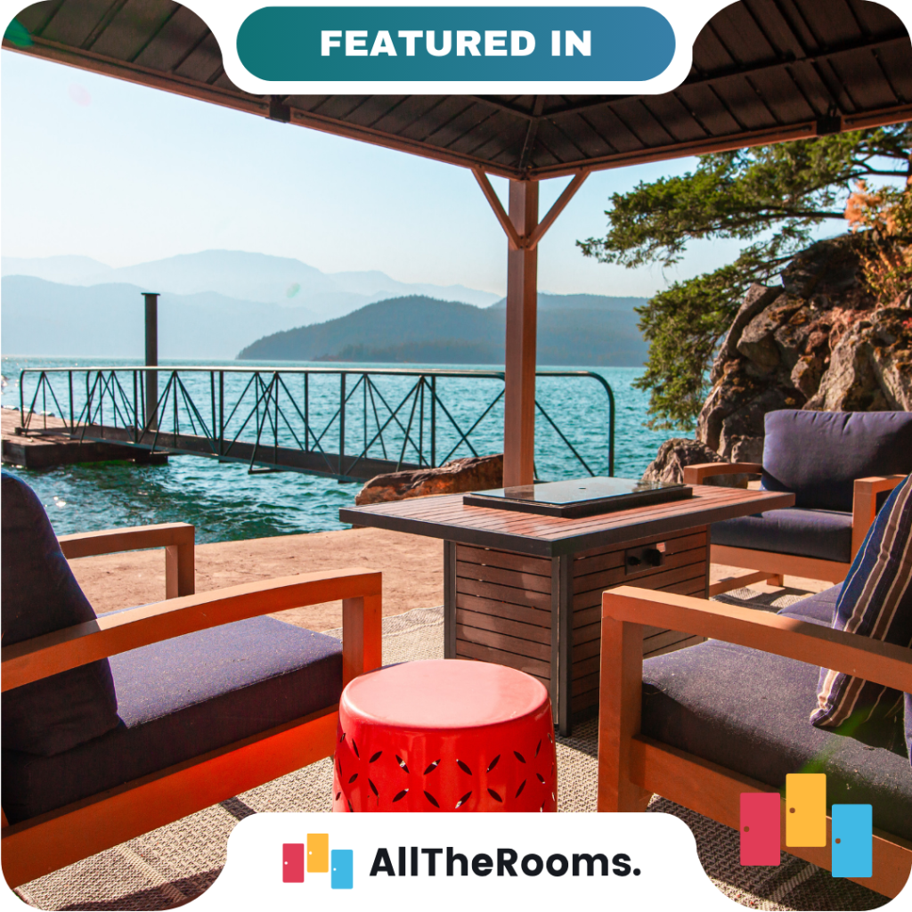 Outdoor patio with cushioned chairs, a small fire table, and a red stool overlooking a lake and mountains, under a shaded pergola near a dock—perfect for relaxing with friends or partners.